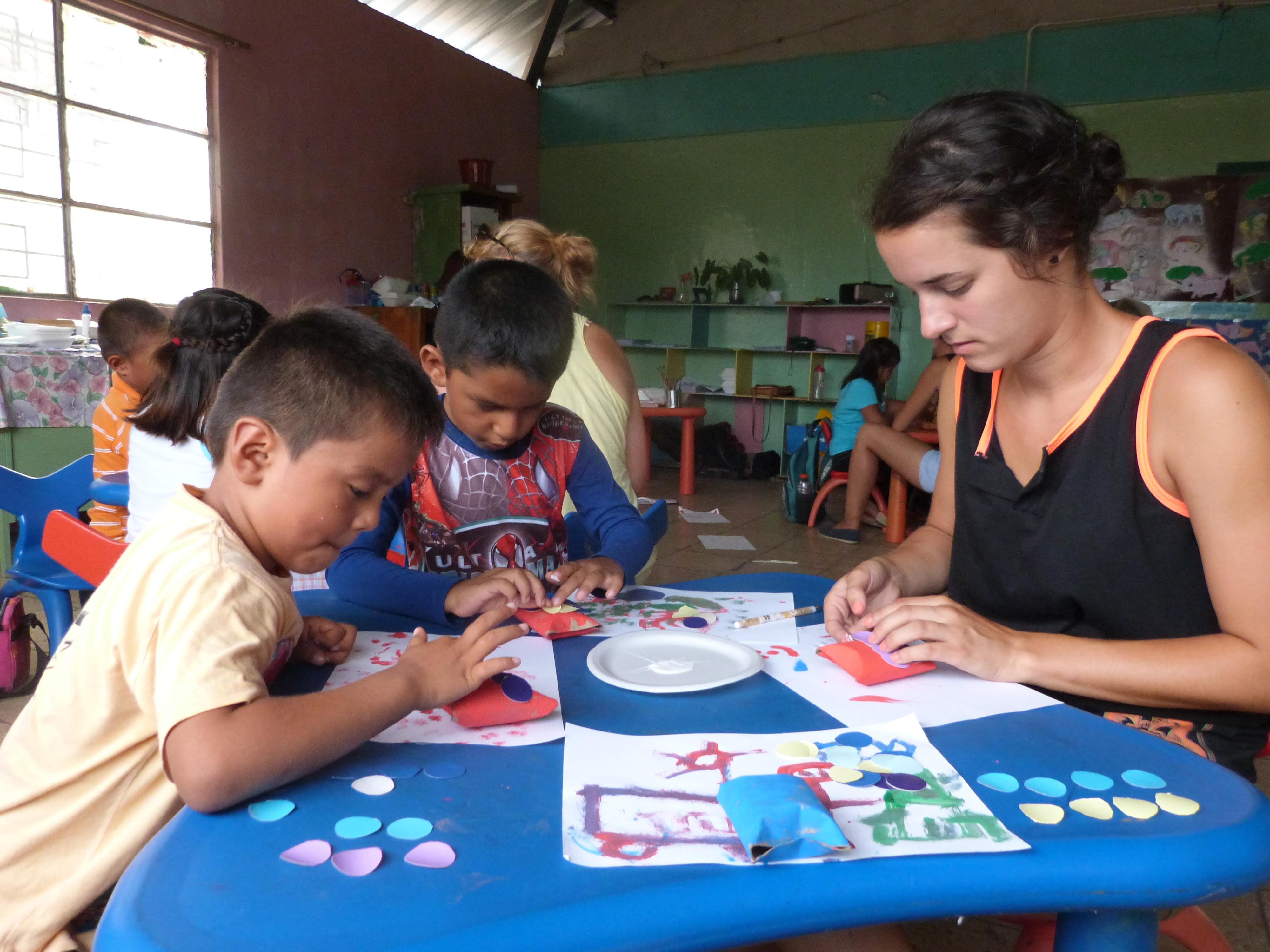 Female Childcare volunteer helps two children with arts and crafts in a day care centre during a project in Ecuador. Female Projects Abroad volunteer with children in Ecuador is doing crafts during Childcare Project.